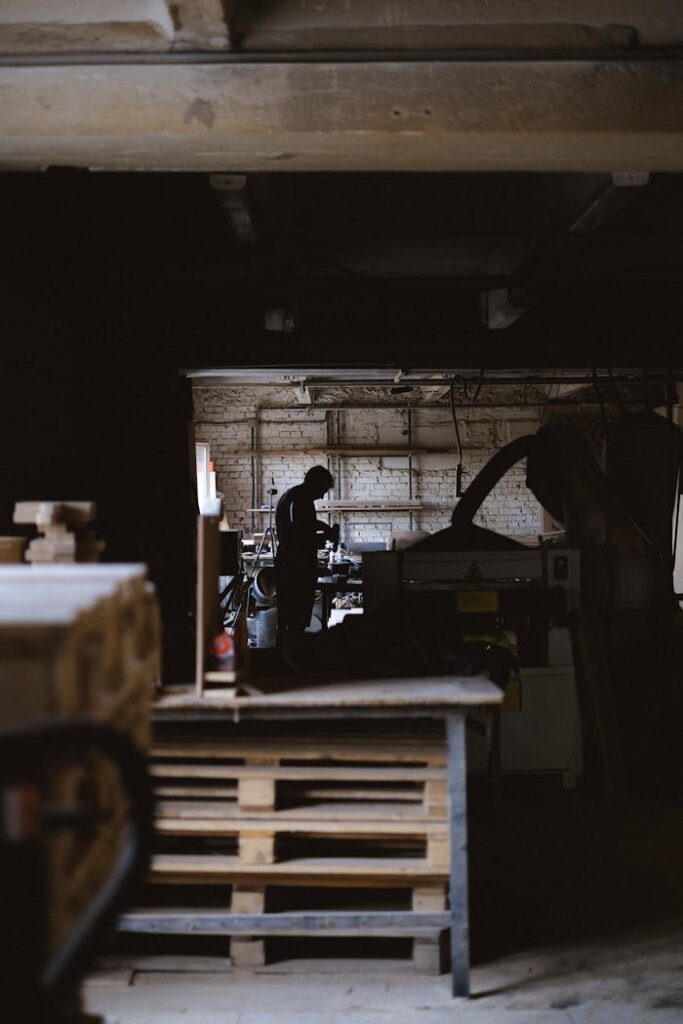 Side view of distant anonymous male artisan standing at special equipment while working with wood in professional carpentry with stair and wooden planks